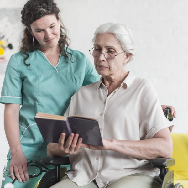 senior-woman-sitting-wheelchair-reading-book-with-nurse-standing